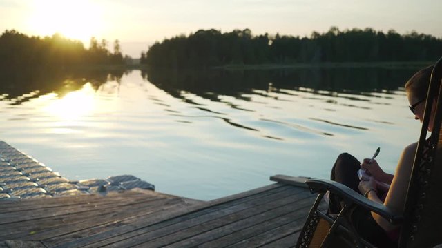 Young white millenial woman with glasses journaling while sitting in chair on pier overlooking beautiful scenic lake vista with trees at sunset