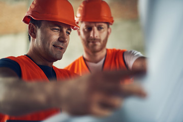 Close up of smiling builder in orange helmet standing near his coworker and smiling while looking...