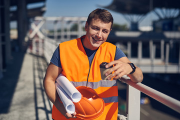 Confident positive young builder wearing an orange vest and smiling while standing alone with coffee and schemes