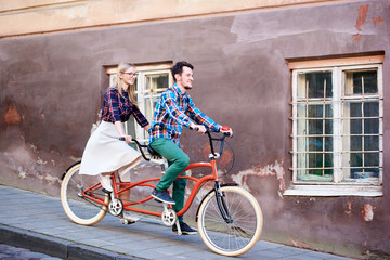 Young handsome man and pretty blond woman riding together tandem double bicycle along paved sidewalk on bright sunny autumn day by old buildings with cracked walls and iron lattice on windows.
