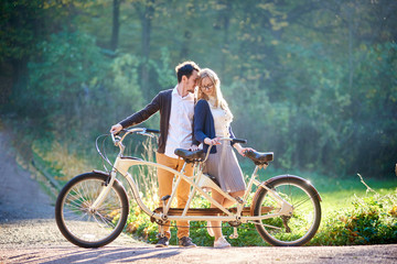 Young happy romantic couple, bearded man and attractive woman close together at tandem double bicycle outdoors in summer park or forest on blurred sunny green trees dense foliage background.