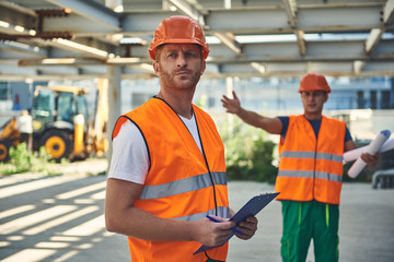 Waist up of two builders in orange form are locating in hangar and holding documents in hands