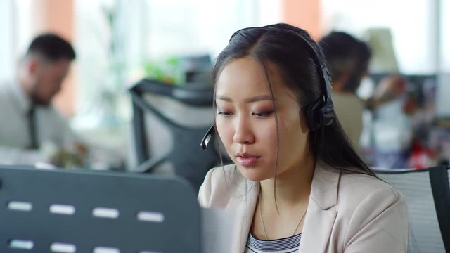 Medium shot of young Asian woman in headset communicating with customer online when working in office