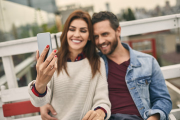 Waist up of happy male and female are sitting on bench and hugging for selfie