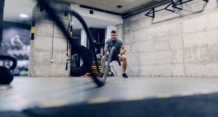 Young Caucasian man in sportswear exercising with battle ropes in a gym.