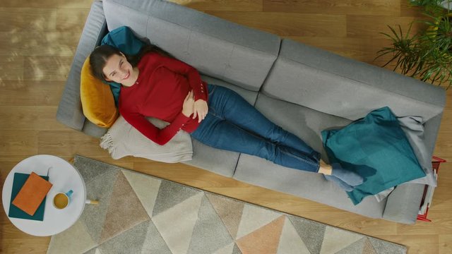 Young Girl in Red Coat and Blue Jeans is Lying Down on a Sofa with Pillows. Looks Above and Smiles. Cozy Living Room with Modern Interior with Carpet, Plants, Coffee Table and Wooden Floor. Top View.