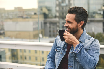 Waist up of happy male in denim jacket is sitting in cozy cafe and drinking coffee. Copy space on left side