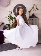 Fototapeta premium little girl in white shiny dress with bow. kid christmas costume. young girl princess in white room is standing from the back like princess in white fancy skirt 