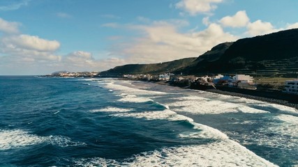 aerial drone image beautiful stunning surf beach with waves and swell coming in in front of a traditional tiny cute village high cliff hills on a sunny day