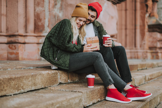Side view portrait of beautiful girl in hat holding gift box while enjoying company of her boyfriend. Bearded man with cup of coffee flirting with lady and smiling