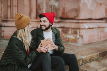 Portrait of charming girl in hat accepting beautiful gift box from her boyfriend. They looking at each other and smiling while sitting on steps