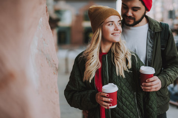 Waist up portrait of charming girl in hat holding cup of coffee while handsome bearded man looking...