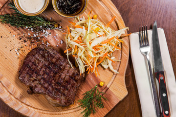 Big meat steak with vegetables, greenery and sauces, knife and fork on wooden table in luxury restaurant