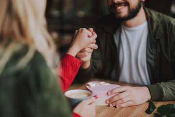 Close up of young gentleman holding hand of young woman while sitting at the table with pink gift box