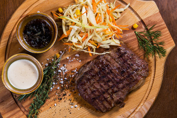 Big meat steak with vegetables, greenery and sauces on wooden table in luxury restaurant