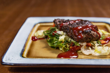 Big meat steak with vegetables, greenery and sauces in square plate on wooden table in luxury restaurant