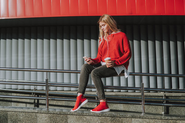 Full length portrait of charming girl in earphones holding cup of coffee and smartphone while...