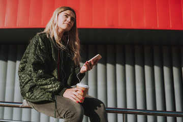 Portrait of charming girl in earphones holding cup of coffee and smartphone while enjoying favorite...