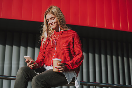 Portrait of charming girl in earphones holding cup of coffee and smartphone while sitting on handrail. She is looking at phone display and smiling