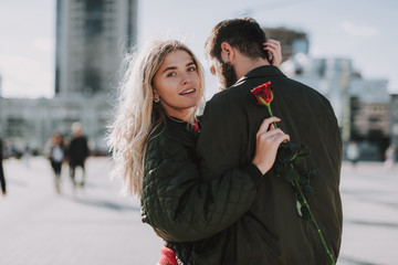 Waist up portrait of charming blond girl holding red rose and tenderly embracing her man. She is looking at camera and smiling
