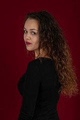 Naklejka premium Waist up portrait of a young woman wearing black dress with long curly hair on a dark burgundy background in the studio