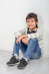 Studio shot of a young laughing boy wearing a white shirt and jeans with headphones siting on the floor by the wall