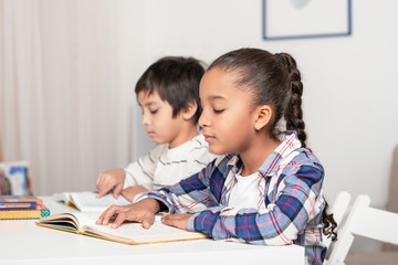 Children, boy and mullata girl, sit at the table and read books or textbooks in indoor