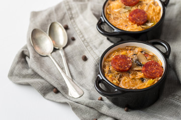 Slovak Christmas national cabbage soup in two small black pots with sausage on the tablecloth background.