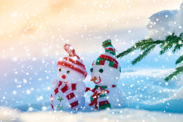 Two small funny toys baby snowman in knitted hats and scarves in deep snow outdoors near pine tree branch. Happy New Year and Merry Christmas greeting card.