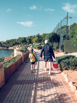 Back view aged grey male in sportswear with pail and little child with blue fillet and tennis racket going on footpath near water in sunny day