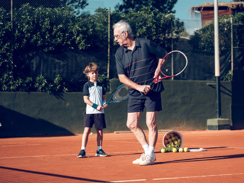 Old man and boy with tennis rackets near bucket with balls on hard court