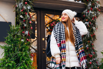 girl in winter clothes and a backpack cute smiles on the background of a cafe with glass doors and Christmas decorations