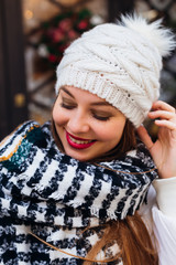 Close-up of a girl with a beautiful makeup with a warm scarf around her neck and a hat with a pompon looking down and smiling against the backdrop of the building.