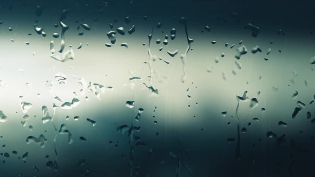 Focus on digitally generated raindrops that fall on a foggy window during the day when it rains and the background is blurred.