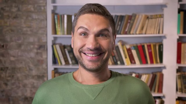 Adult man looking forward laughs and smiles. Bookshelves on the background