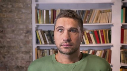 Adult man gets scared and frightened lokking forward. Bookshelves on the background