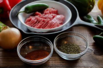 Raw ground beef inside skillet with peppers and other cooking ingredience around with wooden background side view