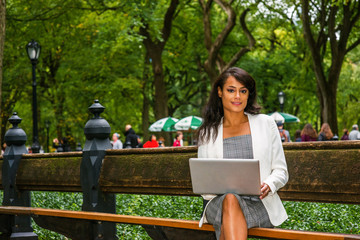 Naklejka premium Young American Woman traveling, working in New York, wearing gray patterned open neck dress, white coat, sitting on long bench at park under green trees, working on laptop computer, looking, thinking