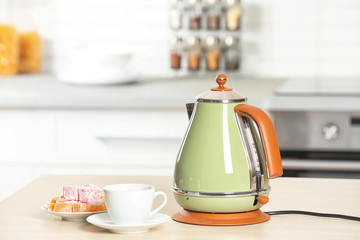 Stylish electrical kettle, cup and plate with sweets on table against blurred room interior. Space for text