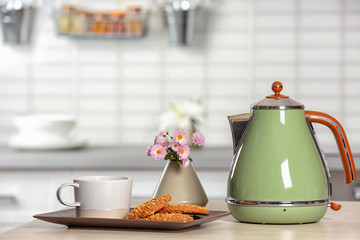 Stylish electrical kettle, cup and cookies on table against blurred room interior. Space for text