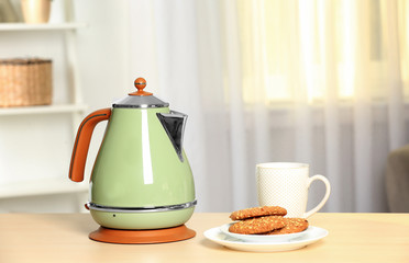 Stylish electrical kettle, cup and plate with cookies on table against blurred room interior. Space for text