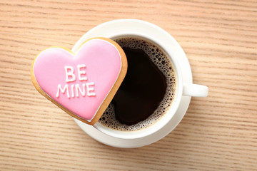 Heart shaped cookie with written phrase Be Mine and cup of coffee on wooden background, top view....