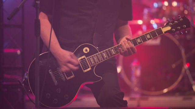 Close up scene of electric guitar player on performing stage during playing solo on concert with colourful scenic illumination via powerful lights. Unicolor simple photography