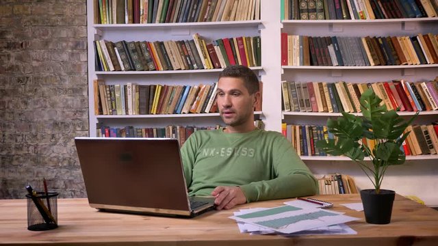 Irritated businessman works on the laptop and makes negative comments. Bookshelves on the background