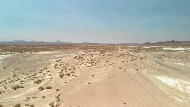 Aerial Footage of a desert with small shrubs and a dirt road. There are brown mountains in the horizon 