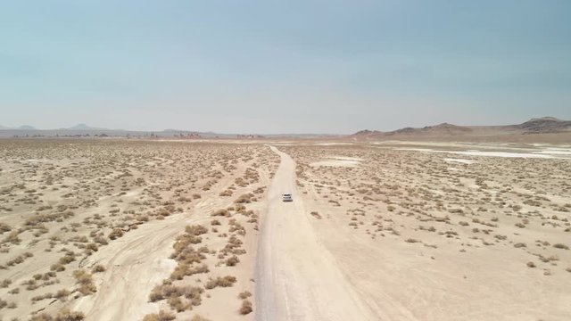 Aerial following shot of a silver car driving on a dirt road in a desert. 