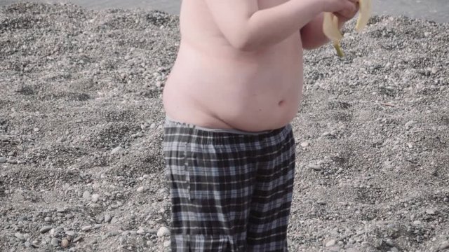Boy with overweight on the beach eating a banana. Slow motion