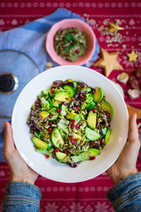 Hands holds big bowl fresh green salad with avocado, spinach, cucumbers, pomegranate grains. Vegan vegetarian Christmas lunch, dinner food on red tablecloth