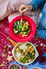 Christmas lunch, dinner food. Baked, roasted or grilled vegetables in cast iron skillet. Brussels sprout, cauliflower. Woman hands eat, take food from red plate or bowl. Hold on legs