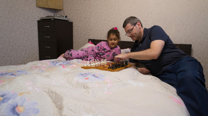 Little girl playing chess with her dad at home, lying on a soft bed wide shot